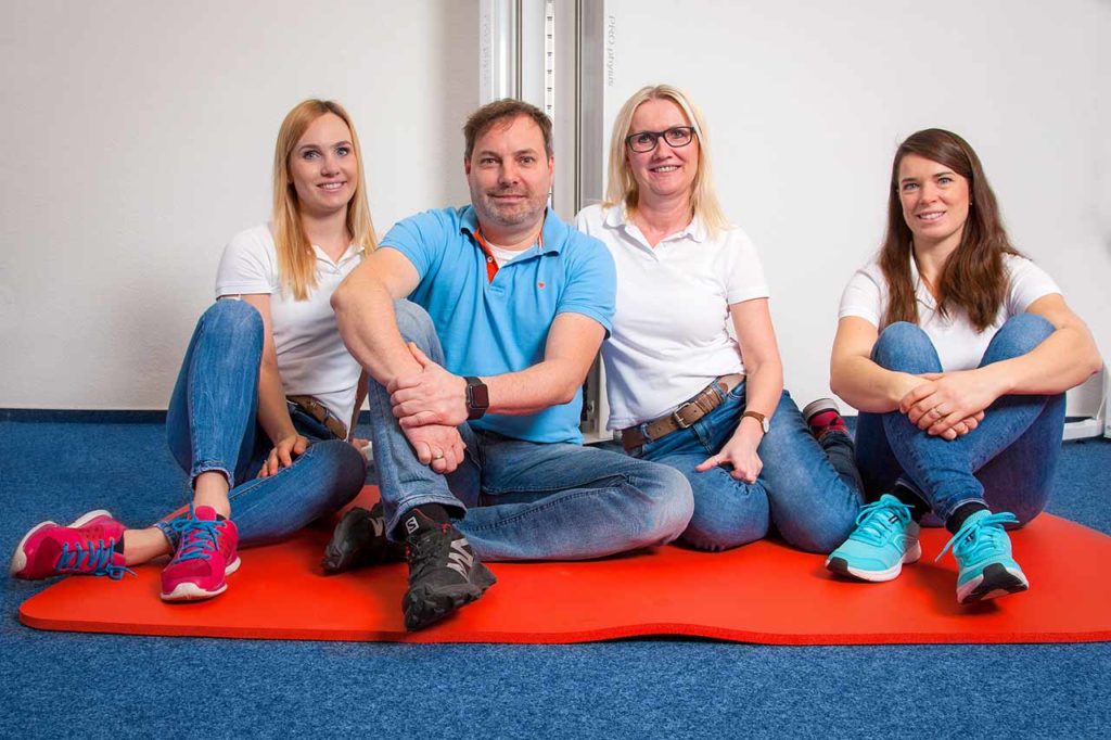 Das Team von Pawel's Physio, der Praxis für Physiotherapie in Witten, sitzend auf einer roten Decke im Behandlungszimmer.
